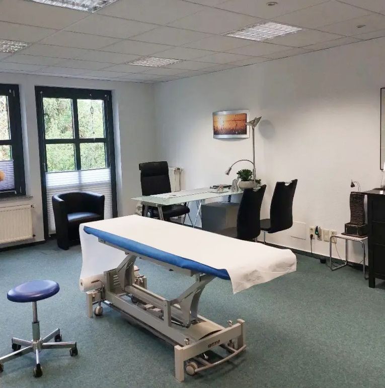 Ein medizinischer Untersuchungsraum mit einem verstellbaren Bett, einem Schreibtisch mit Stühlen und einem Fenster mit Bäumen draußen.